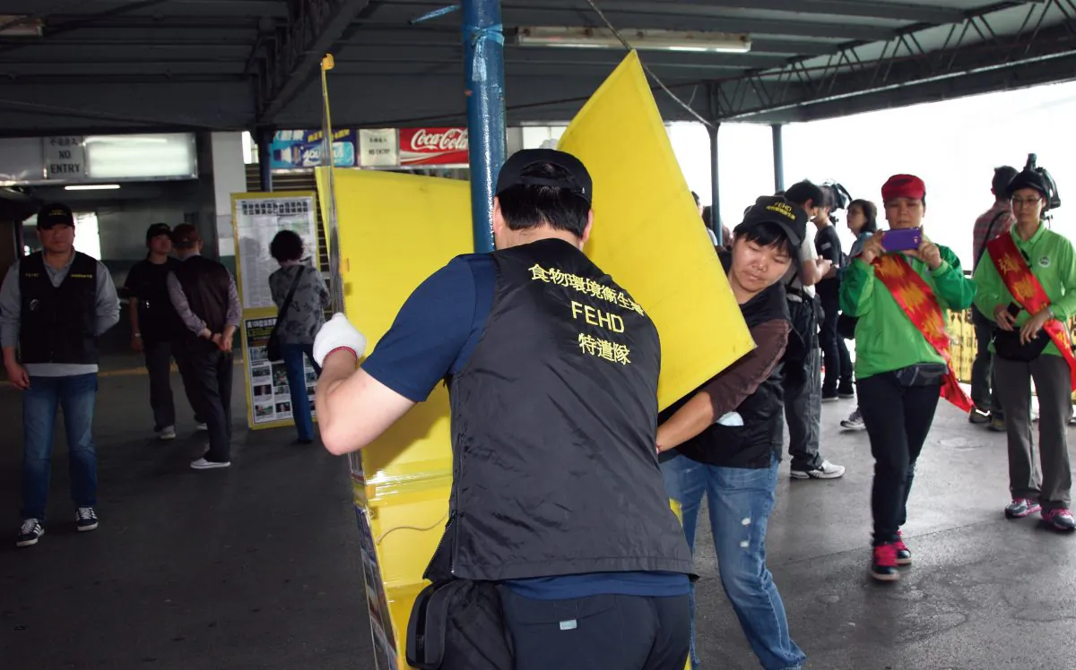 Individuals not wearing the regular uniform of the Hong Kong Food and Hygiene Department are seen removing Falun Gong materials from the information site in the neighborhood of Tsim Sha Tsui, on April 15. In the background to the right, the two individuals wearing green shirts and sashes are members of the Hong Kong Youth Care Association. In line with Epoch Times policy of not publishing hate speech, the words on their sashes, which repeat Communist Party propaganda attacking Falun Gong, have been blurred. (Epoch Times) 
