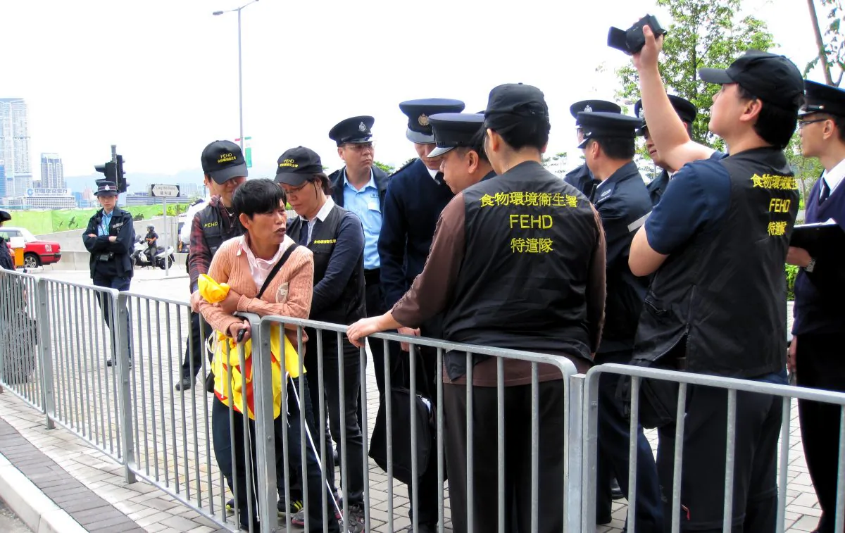 A Falun Gong practitioner is surrounded by members of the Food and Environmental Hygiene Department, along with police, outside the Central Government Complex at Tamar in Hong Kong on May 2. The authorities arrested the practitioner, an unusual action, after she refused to yield a fourth banner. (Bill Cox/The Epoch Times)