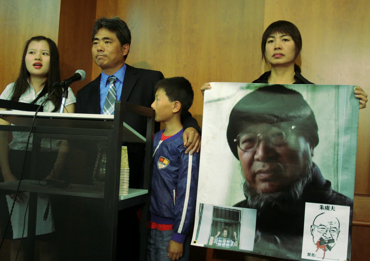 Zhu Qiaofu with son speaks at a news conference to call for the release from prison of his brother Zhu Yufu. Their sister Zhu Xiaoyan holds up photo of Zhu Yufu, who is serving a seven year sentence for writing a poem. Person on the far left is a translator. News conference took place June 6. (Gary Feuerberg/ The Epoch Times)