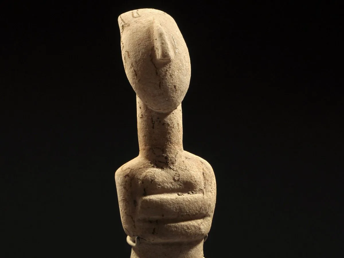A detail from an elegant stylized nude figure of the early Spedos type, her arms are folded beneath her breasts, her slender lyre-shaped head with wedge-shaped nose and long oval neck; incised details, with traces of red pigment and strong root marks. Early Bronze Age II, ca. 2600-2500 BC. H. 9 11/16 in. (24.6 cm.) (Royal-Athena Galleries)