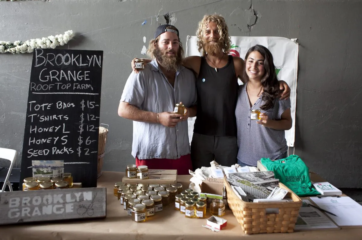 (L-R) Beekeeper Asher landes, Beekeeper Mike Densham and Program manager at City Growers Nicki Sherman pose in front of the Brooklyn Grange Rooftop Farm booth on the High Line, July 31. (Samira Bouaou/Epoch Times)