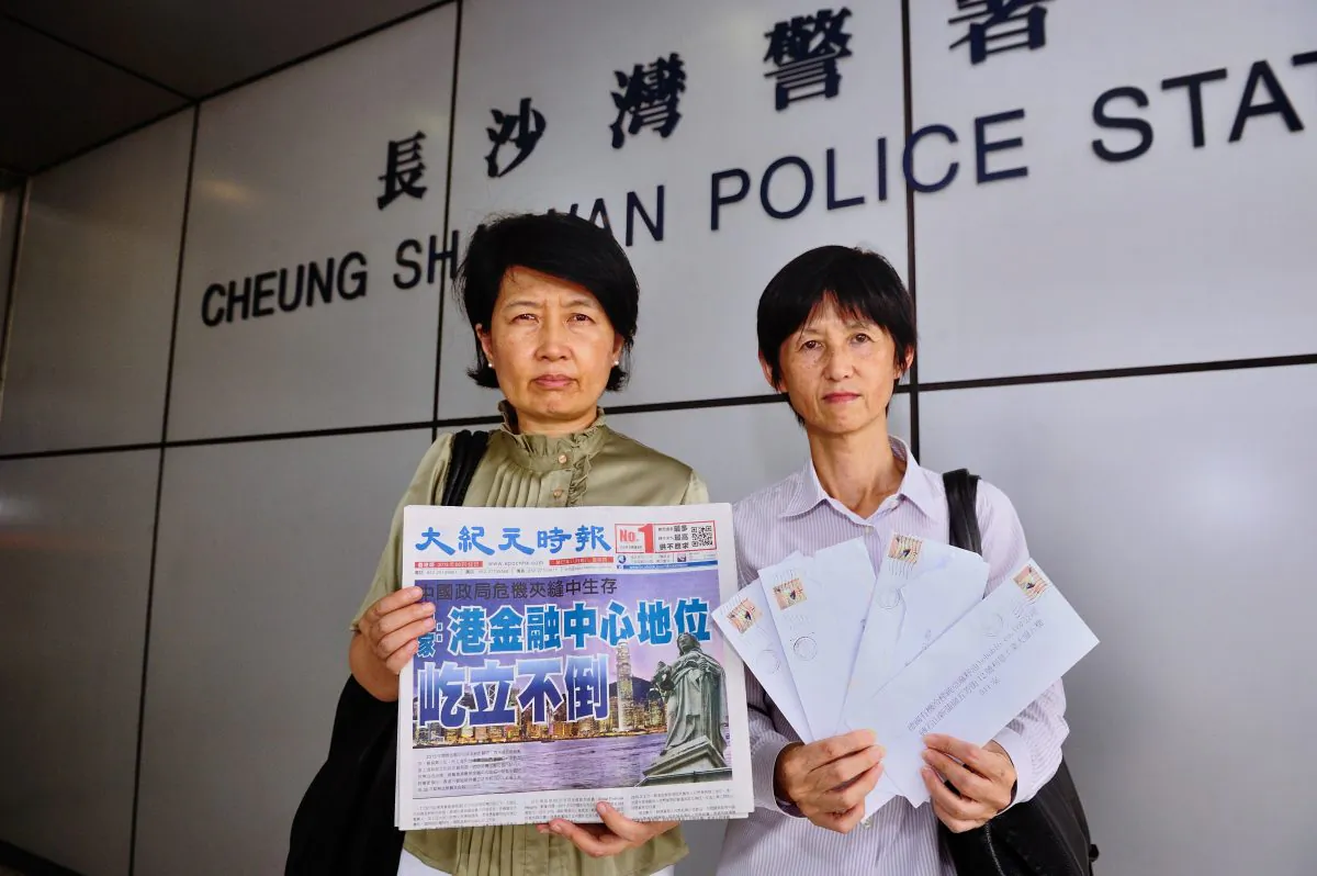 Two staff members of the Chinese-language edition of the Hong Kong Epoch Times stand outside the police station where they reported a campaign of harassment against the paper, on Sept. 12. The woman on the left holds a copy of the day's paper, and the woman on the right holds letters sent to advertisers that were meant to discredit the paper. (Song Xianglong/Epoch Times) 