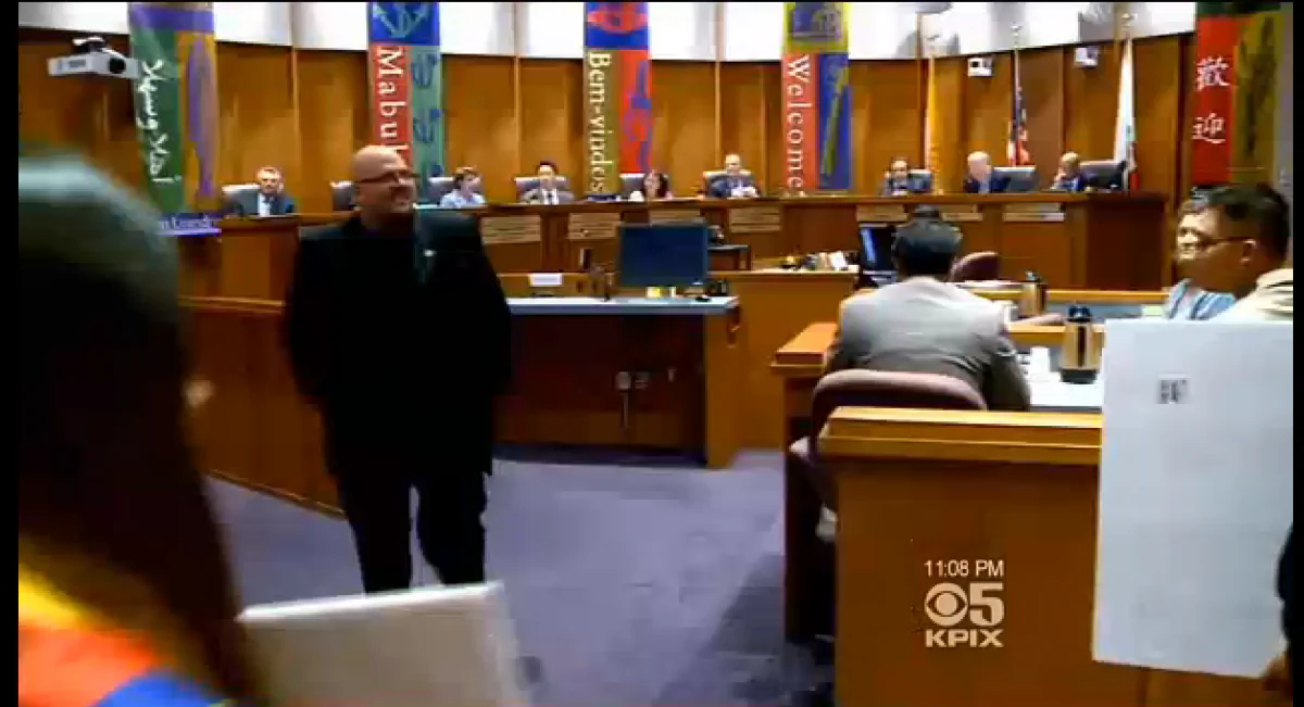 A Tibetan protester (left) walks into the City Council meeting on Sept. 16 to speak against the proposal that the flag of the People's Republic of China be raised on Oct. 1, the PRC's National Day. The proposal has met with strenuous objections. (Screenshot/KPIX/Epoch Times)
