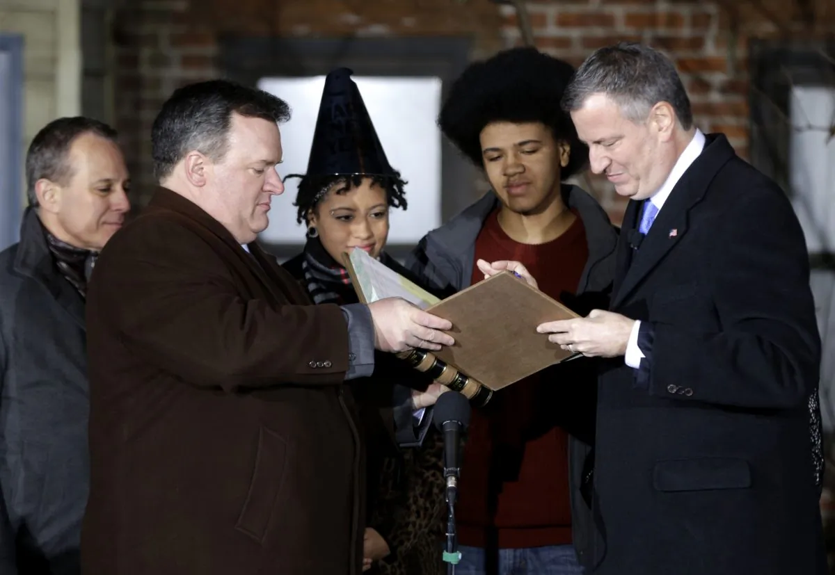 City Clerk Michael McSweeney (2nd L) assists Bill de Blasio (R), as he signs the oath of office while (L–R) State Attorney General Eric Schneiderman, Chiara de Blasio, and Dante de Blasio look, New York, Jan. 1, 2014. De Blasio took the oath of office moments after midnight at his home in Park Slope, Brooklyn, his inauguration will be celebrated at noon on the steps of City Hall when he takes the oath again, which will be administered by President Bill Clinton. (AP Photo/Seth Wenig, Pool)