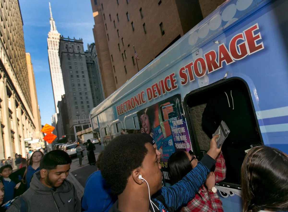 Students pay a dollar to check their electronic devices at a van, before school, adjacent from the Norman Thomas High School building in New York on Thursday, Oct. 9, 2014. New York City is preparing to end its longtime ban on cellphones in public schools, fulfilling a campaign promise of Mayor Bill de Blasio's but signaling the death knell for an industry that sprung up near dozens of schools where teens could park their phones in a van for a dollar a day. (AP Photo/Richard Drew)
