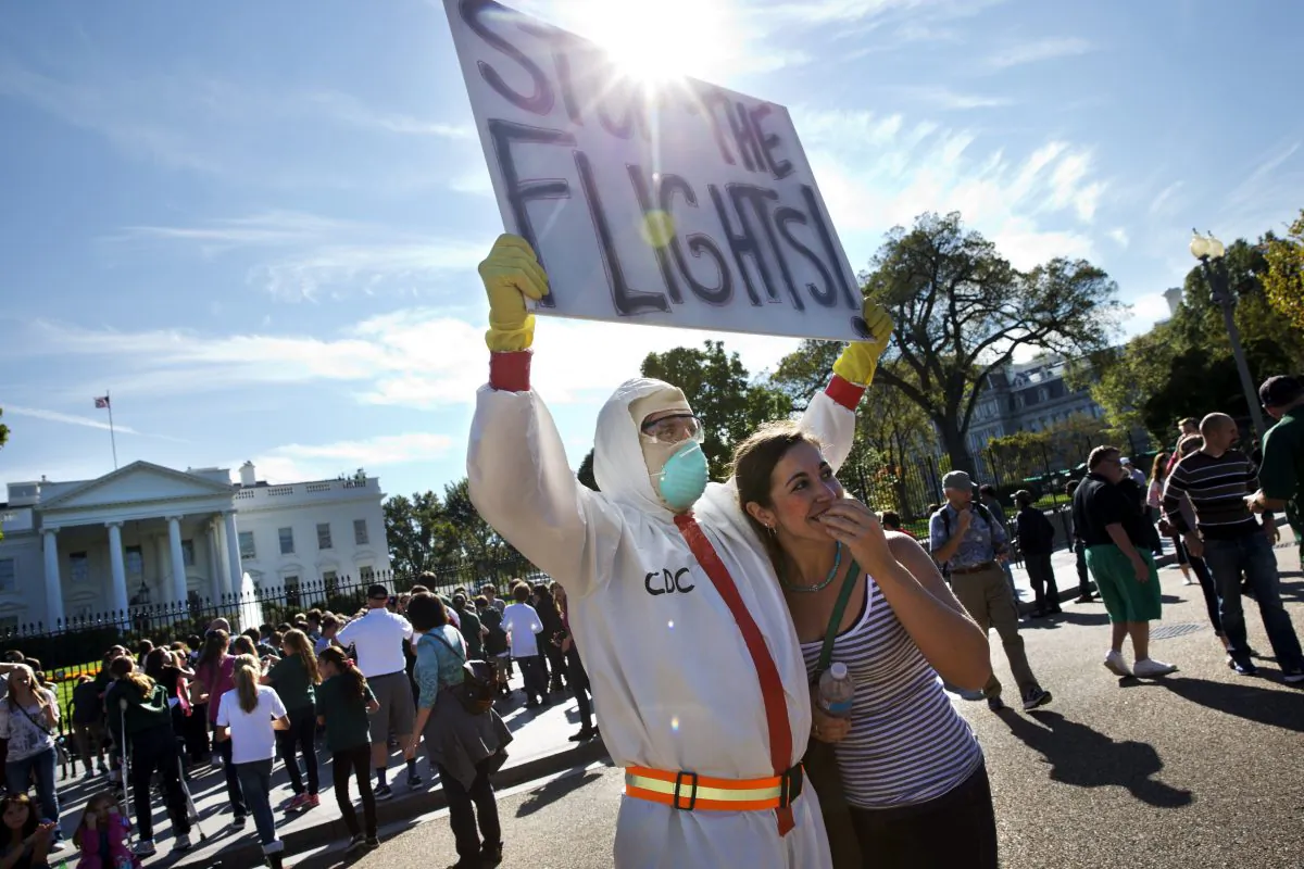  Jeff Hulbert, left, of Annapolis, Md., protests U.S. handling of Ebola cases, as Mary Wills, right, of Cleveland, playfully covers her face while taking a photo with Hulbert, outside of the White House. A ban on travel from West Africa might seem like a simple and smart response to the frightening Ebola outbreak there. It’s become a central demand of Republicans on Capitol Hill and some Democrats, and is popular with the public. But health experts are nearly unanimous in saying it’s a bad idea that could backfire. (AP Photo/Jacquelyn Martin)