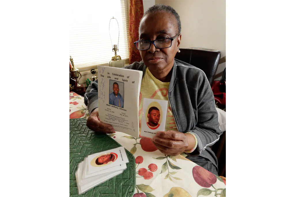 June Broer displays a funeral program and a prayer card featuring images of her only son, Mark Johnson, in Paterson, N.J., Oct. 18, 2014. For days Johnson demanded medical care from jail guards at Rikers Island as he continued to have bloody stools before he died of a bacterial infection in his stomach and small bowel. (AP Photo/Julio Cortez)