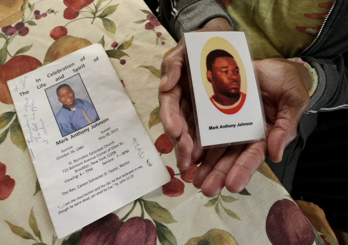 June Broer displays a funeral program and a prayer card featuring images of her only son, Mark Johnson, in Paterson, N.J., on Oct. 18, 2014. For days, Johnson demanded medical care from jail guards at Rikers Island as he continued to have bloody stools before he died of a bacterial infection. (AP Photo/Julio Cortez)