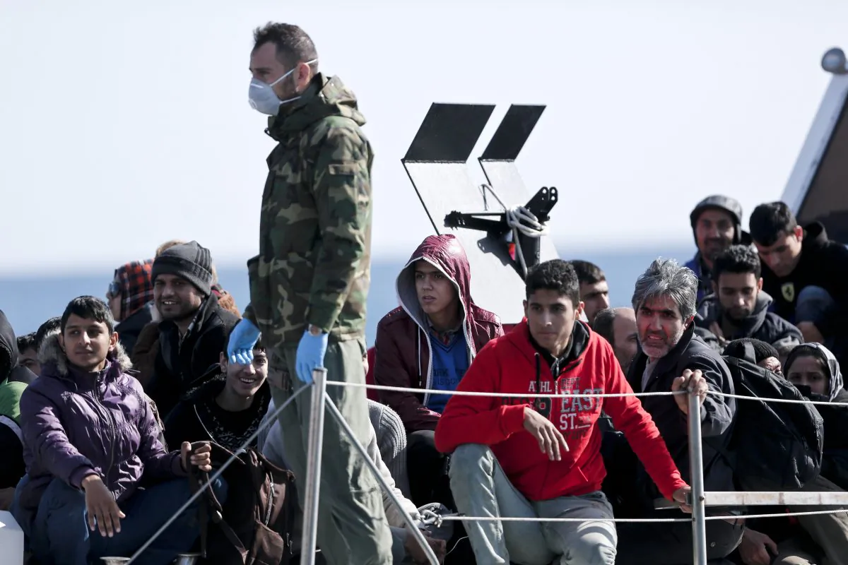 A Greek coast guard officer, wearing a mask for fear of infectious disease, stands next to a small group of immigrants, on a launch carrying them to shore from a crippled smuggling ship with more than 600 people, at the coastal Cretan port of Ierapetra, Greece, on Thursday, Nov. 27, 2014. The ship, whose passengers are mostly Syrians,  including children, women and elderly men, suffered engine failure 70 nautical miles off Ierapetra on Tuesday. The migrants will be given temporary shelter at an Ierapetra indoor basketball stadium. (AP Photo/Petros Giannakouris)
