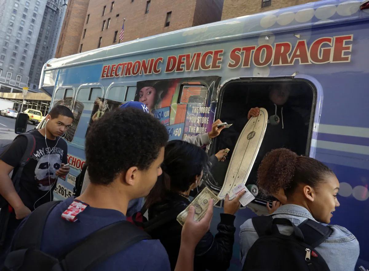 Students from the adjacent Norman Thomas High School building, pay a dollar to check their electronic devices, at a van in New York City on Thursday, Oct. 9, 2014. The decades-long ban on cell phones is to be lifted in NYC schools in two months. (AP Photo/Richard Drew)