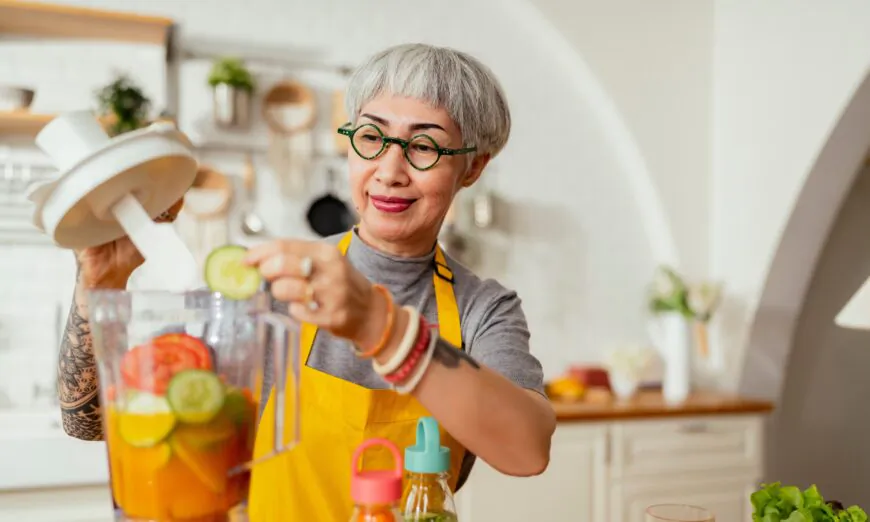 Mature smiling tattoo woman eating salad, fruits and vegetables. Attractive mature woman with fresh green fruit salad at home. Senior woman apron standing in the kitchen counter relaxing in house