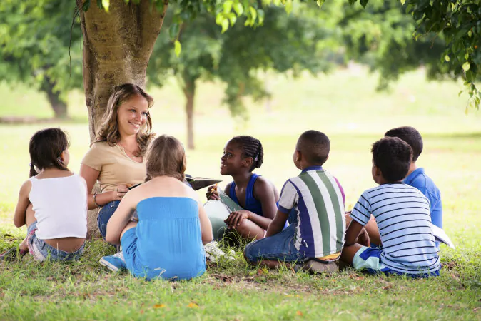 A young woman at work as educator reading book to boys and girls in the park via Shutterstock*