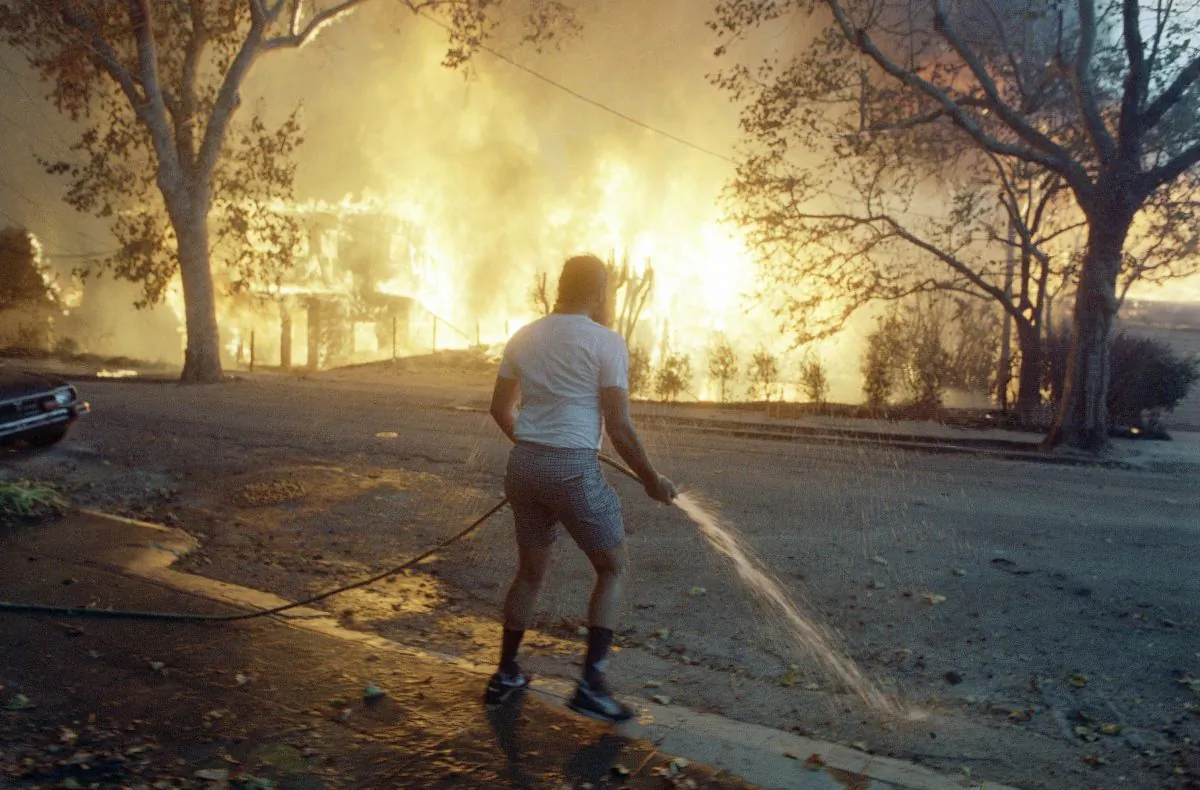In this Sunday, Oct. 20, 1991, photo, Berkeley resident Jim Beatty stands with a garden hose as homes burn out of control in the Berkeley and Oakland hills area on in Berkeley, Calif. (AP Photo/Kevin Rice, File)