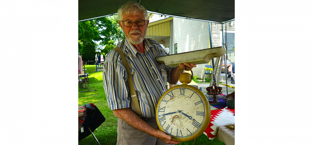 Collector, dealer, and antique show promoter Bill Dobson at his booth at the Perth Antique Show and Sale on July 4, 2015 in Perth, Ont. He is holding a wooden sign shaped like a watch that would have been hung outside a shop to indicate watch or clock repairs. (Pam McLennan/Epoch Times)