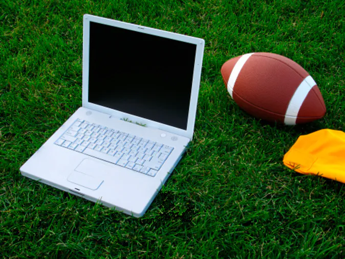 This color photo shows a closeup view of a white laptop computer, a football, and a bright-yellow penalty flag on a deep-green grassy football field in an attempt to create a fantasy-football concept image. The grassy turf fills the entire background of the image.