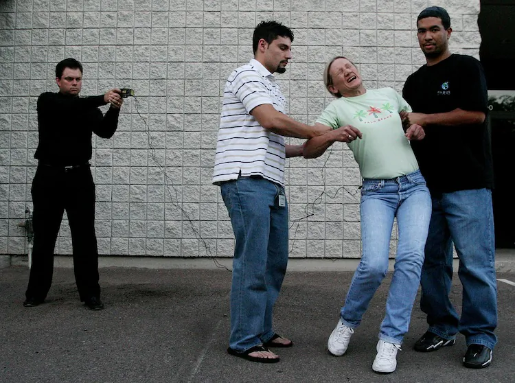 TASER International employee Evelina Self is held by employees Eric Rivera (L) and Charles Foster (R) as Taser co-founder Tom Smith (far left) demonstrats the X26 Taser at the company's headquarters in Scottsdale, Arizona, on July 9, 2004.  (Jeff Topping/Getty Images)