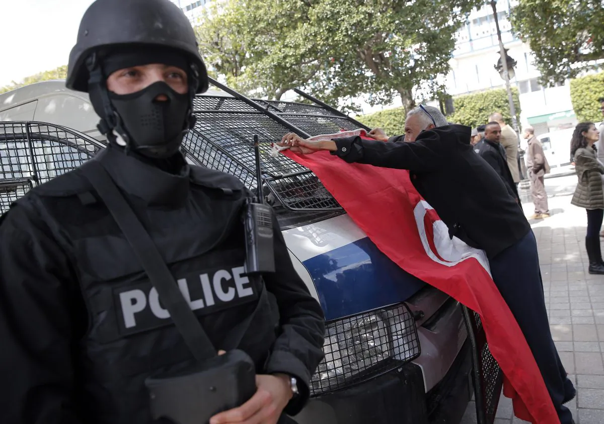 FILE - In this March 20, 2015 file photo, a man places a Tunisian flag onto a police car as a policeman stands guard during a demonstration two days after gunmen attacked the museum and killed scores of people in Tunis, Tunisia.  Seven men spent a week in prison on terrorism charges, suffering what they claim was torture under custody, before a judge released them for lack of evidence. But as they stepped out of the courthouse in early August, plainclothes policemen swooped in and spirited them away. (AP Photo/Christophe Ena, File)