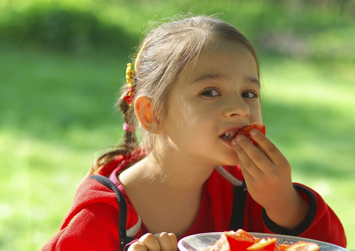 The little girl sits at table in garden and eats tomato. (-101PHOTO-/iStock)
