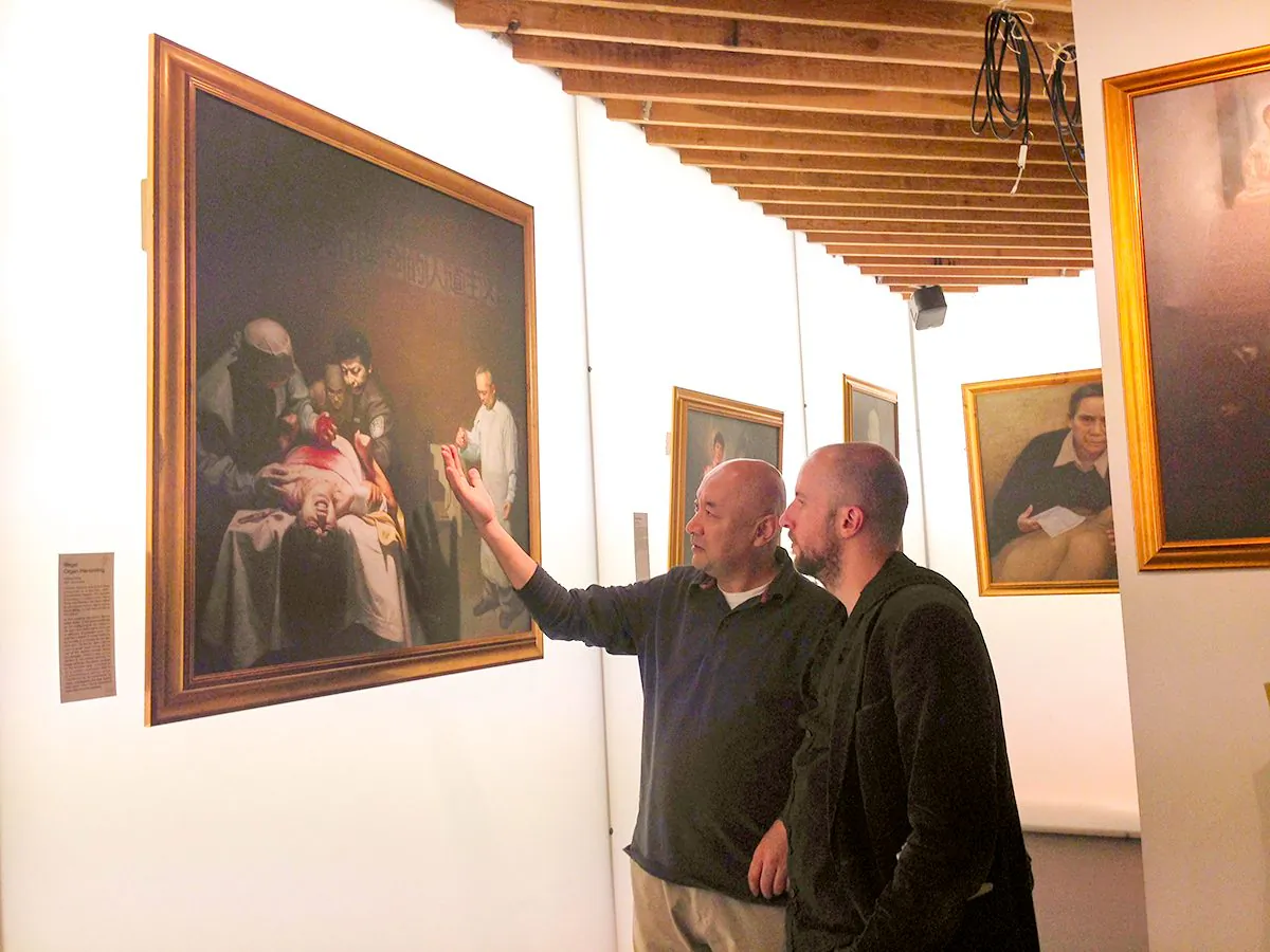 Enver Tohti (left), a former cancer surgeon in China, stands in front of the painting “Illegal Organ Harvesting” on the opening night of the Art of Zhen Shan Ren International art exhibition at the Proud Archivist, London, on Sept. 1, 2015. (Xiaomin Pang)