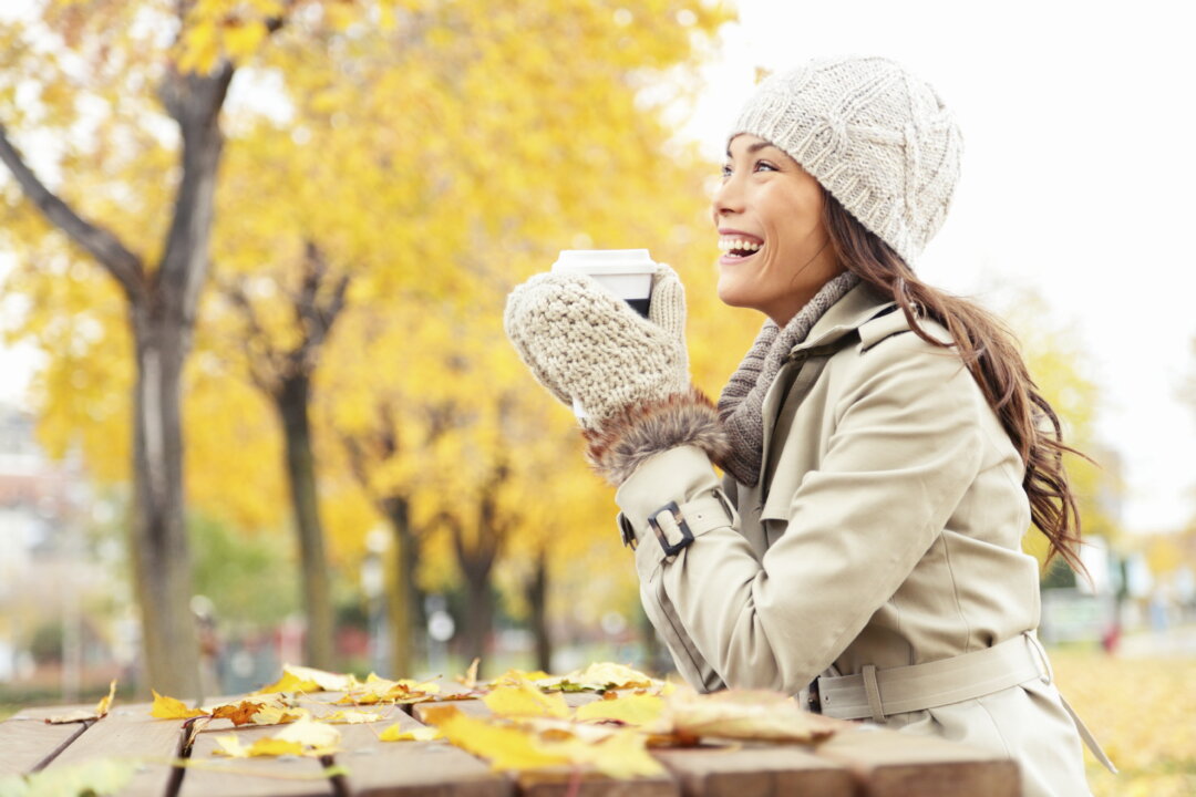 Fall concept - autumn woman drinking coffee on park bench under fall foliage. Beautiful young modern woman smiling happy and cheerful in trench coat.
