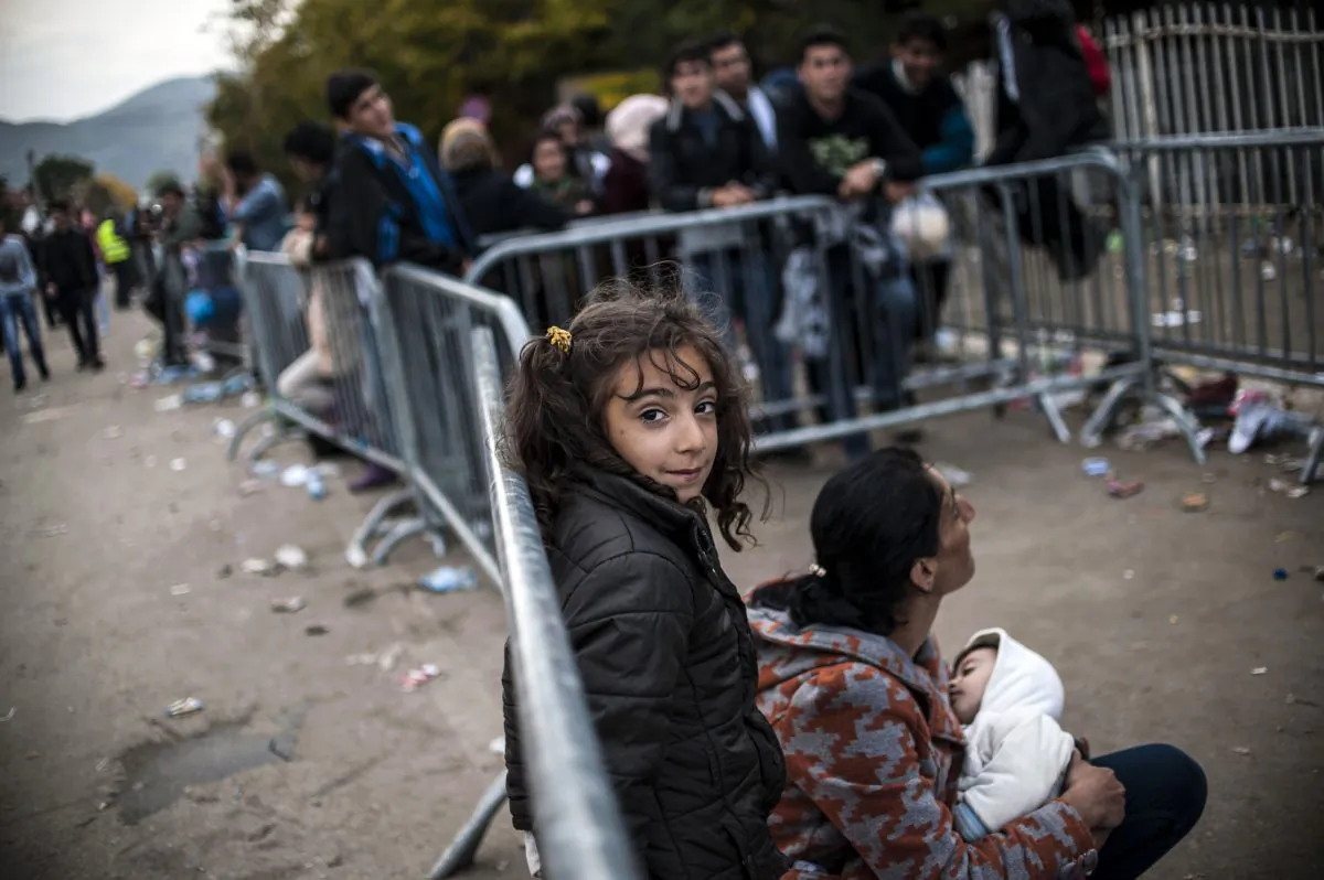 A young girl stands smiles from behind a barrier as migrants and refugees wait to be registered at a refugee center in Presevo, southern Serbia, on October 19, 2015. (ARMEND NIMANI/AFP/Getty Images)