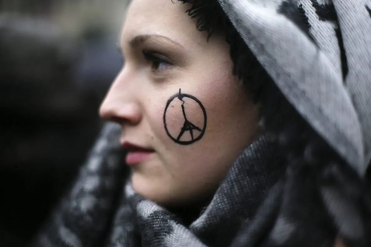 A woman has a peace sign combined with the Eiffel Tower painted on her face as she arrives for a minute of silence for the victims of Nov. 13 attacks in Paris, in front of the French Embassy in Berlin, on Nov. 15, 2015. (AP Photo/Markus Schreiber)