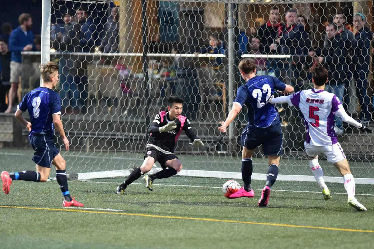 HKFC player James Beacher (Blue), watched by teammate Frederick Schipper, scores in the 22nd minute of the second half in their 1-nil victory over Double Flower at Sports Road on Sunday Jan 31, 2016. (Bill Cox/Epoch Times)