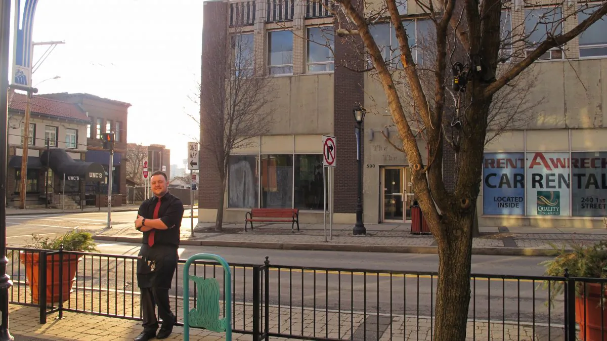 In this Feb. 1, 2016, photo, Eamon Weber poses for a photo stands on the patio of the Wine on Third restaurant where he works in Niagara Falls, N.Y. Across the street, behind him, is the vacant building from which speakers perched on an upper window ledge continually play the University of Iowa fight song every evening. (AP Photo/Carolyn Thompson)