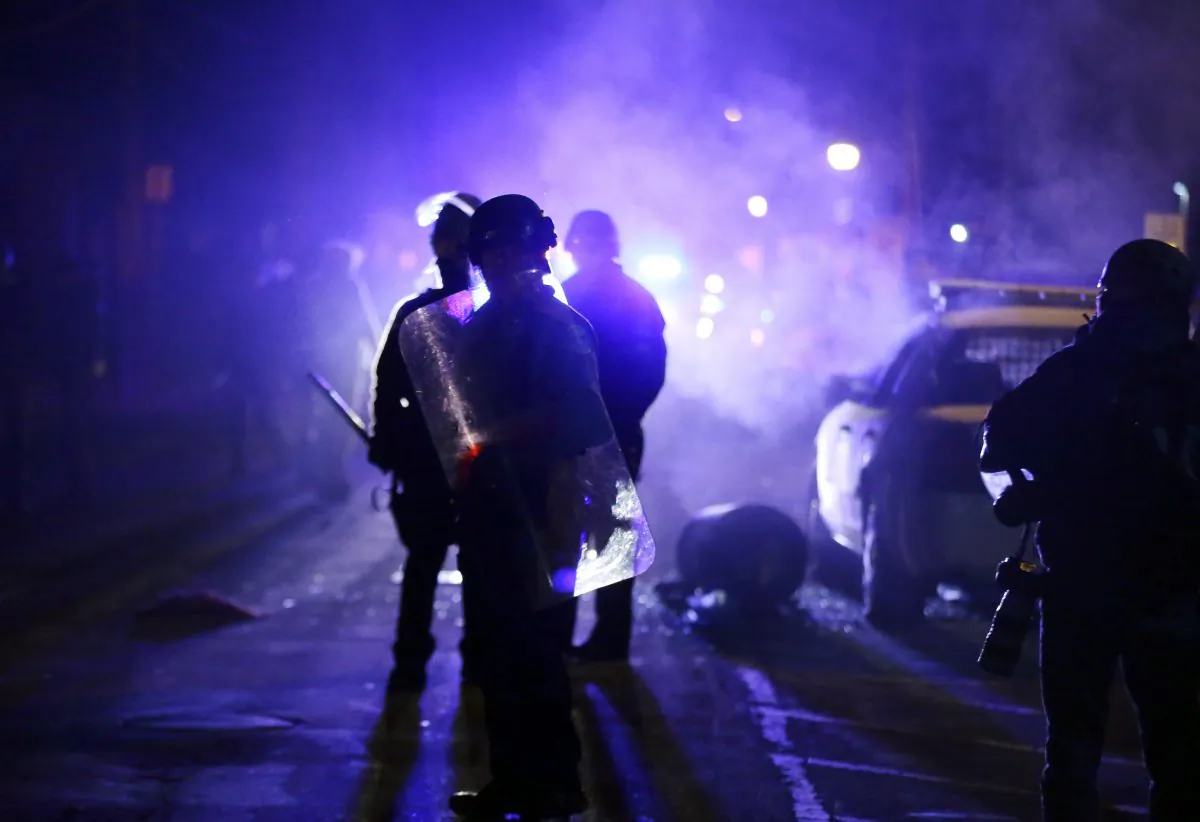 Police officers watch protesters as smoke fills the streets in Ferguson, Mo., on Nov. 25, 2014. In the wake of Michael Brown's death, the federal government wants to require the Ferguson Police Department to check all new hires against a database of police officers stripped of their law enforcement licenses for misconduct. The database contains the names of about 20,000 former officers who have been pushed out of law enforcement. Since it's been used inconsistently, many officers who are stripped of their badges in one jurisdiction are free to move to another. (AP Photo/Charlie Riedel)