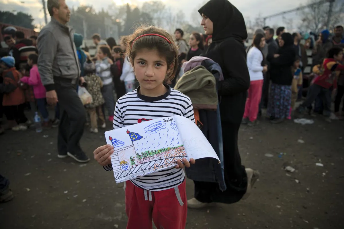 Shaharzad Hassan, 8 year-old from the Syrian city of Aleppo poses with a drawing she made as people wait in line for food rations at the northern Greek border station of Idomeni, Friday, March 11, 2016. At an overcrowded refugee camp on the Greek-Macedonian border, 8-year-old Shaharzad Hassan draws pictures of the harrowing events in her life over the past 18 months: Pictures of death in her home town of Aleppo, Syria, and her perilous journey to Europe. The title, of this drawing seen at top right reads "Famine" .(AP Photo/Vadim Ghirda)