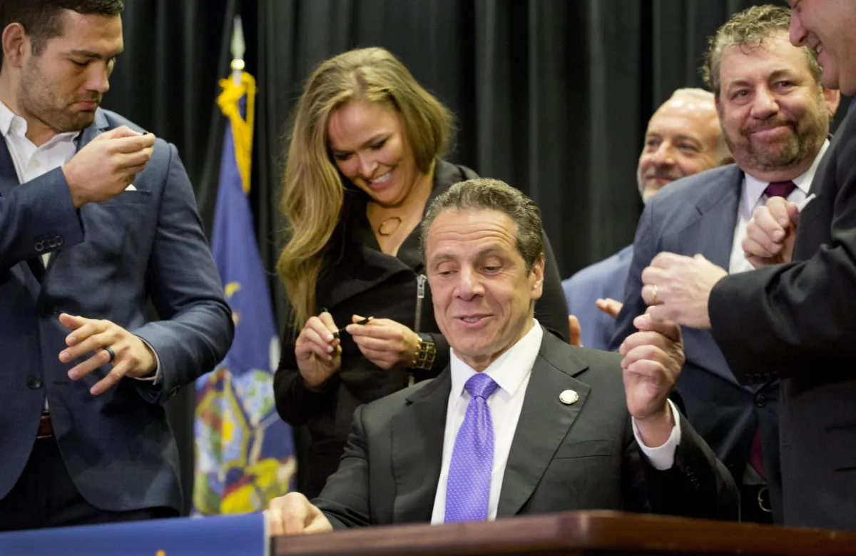 Gov. Andrew Cuomo, center, hands out souvenir pens after signing into law a measure that will allow professional mixed martial arts in New York, Thursday, April 14, 2016. Behind him are UFC athletes Chris Weidman, left, and Ronda Rousey, and James Dolan, executive chairman of Madison Square Garden. New York was the last U.S. state to prohibit the bouts. (AP Photo/Mark Lennihan)