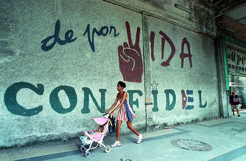 A woman dressed in the colors of the United States flag pushes her baby as they walk past a wall that reads, "A Lifetime with Fidel," June 11, 2001 in Havana, Cuba. (Jorge Rey/Getty Images)