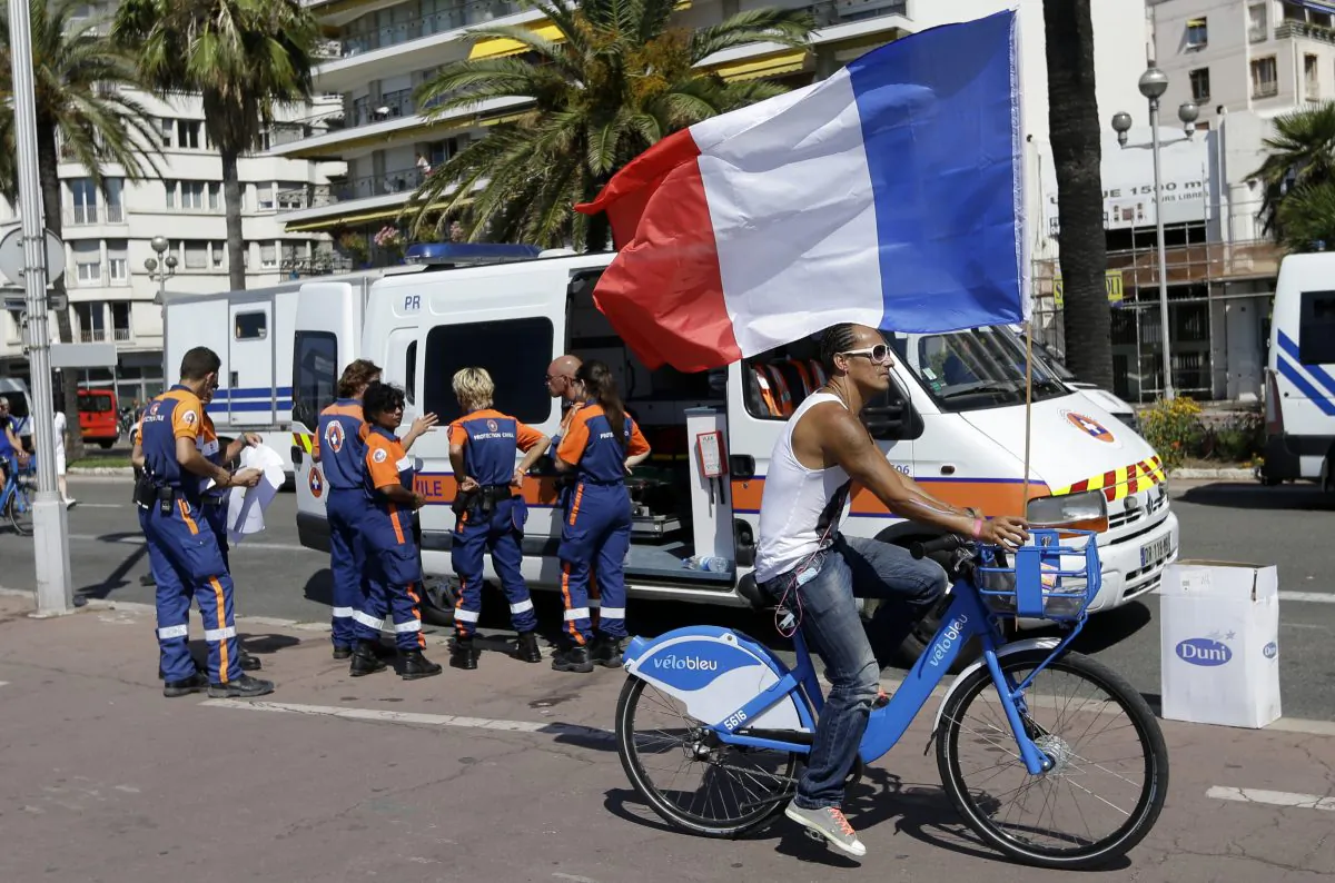 A man rides a bicycle as he holds a French national flag on the Promenade des Anglais in Nice, southern France, Monday, July 18, 2016. France is holding a national moment of silence for 84 people killed by a truck rampage in Nice, and thousands of people are massed on the waterfront promenade where Bastille Day celebrations became a killing field. (AP Photo/Claude Paris)
