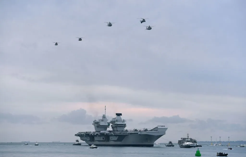 Wildcat and Merlin helicopters fly above the 65,000-tonne British aircraft carrier HMS Queen Elizabeth as tug boats manoeuver it into Portsmouth Harbour in Portsmouth, southern England on Aug. 16, 2017. (Ben Stansall/AFP/Getty Images)
