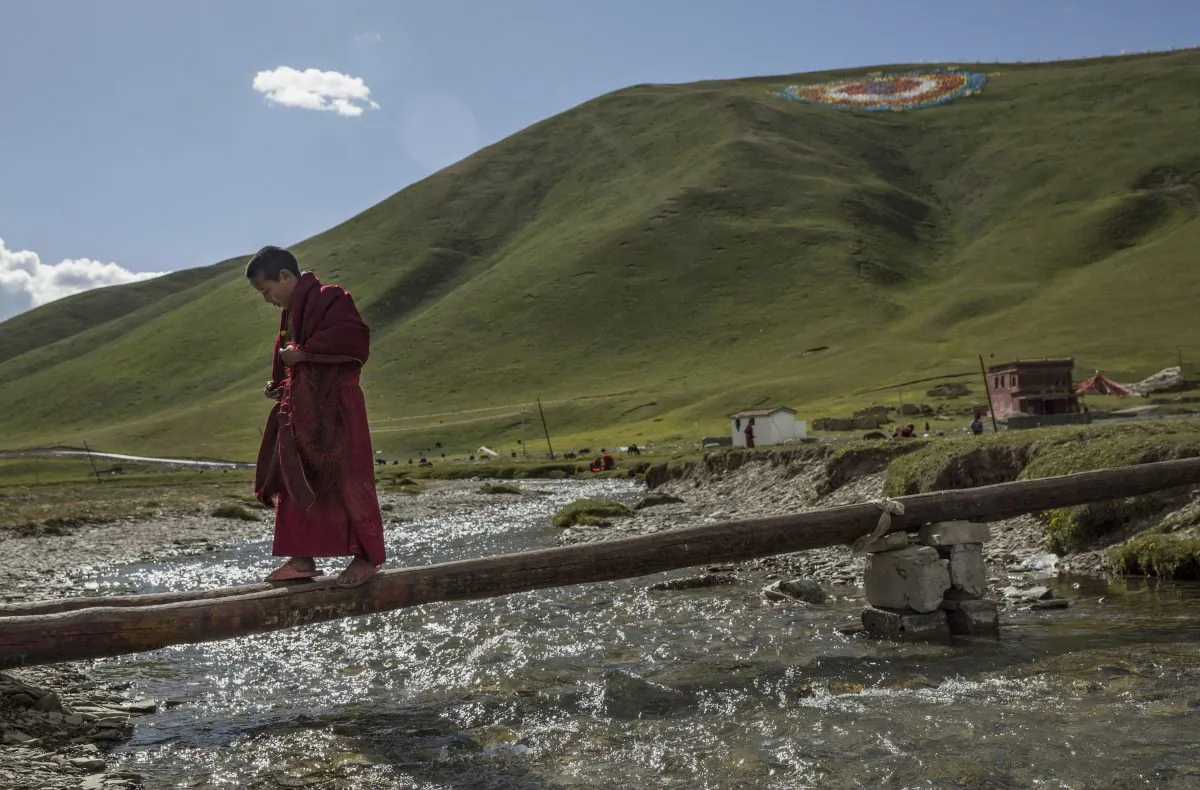 A young Tibetan Buddhist monk walks across a beam over a creek at a monastery next to a government resettlement community for former nomads on the Tibetan Plateau in Yushu County, Qinghai, China, on July 23, 2015. Millions of Tibetans have been forced by the Chinese regime to give up their traditional way of life and resettle in government-designated areas. (Kevin Frayer/Getty Images)