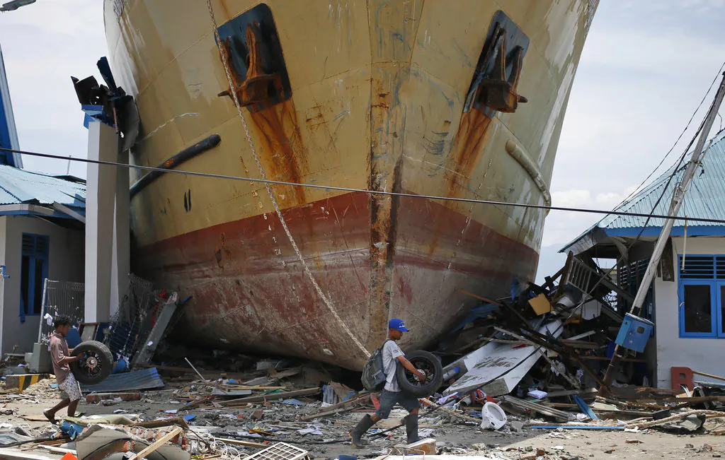 Indonesian men on Oct. 4, 2018, carry car tires past Sabuk Nusantara 39 which was swept ashore by the tsunami in Wani village on the outskirt of Palu, Central Sulawesi, Indonesia. The crew of the hulking ferry dropped by the tsunami in front of a row of houses in an Indonesian village say the wave that drove them onto land was a towering 10-15 meters or higher and came just several minutes after a quake made the ship bounce like a basketball. (AP Photo/Dita Alangkara)