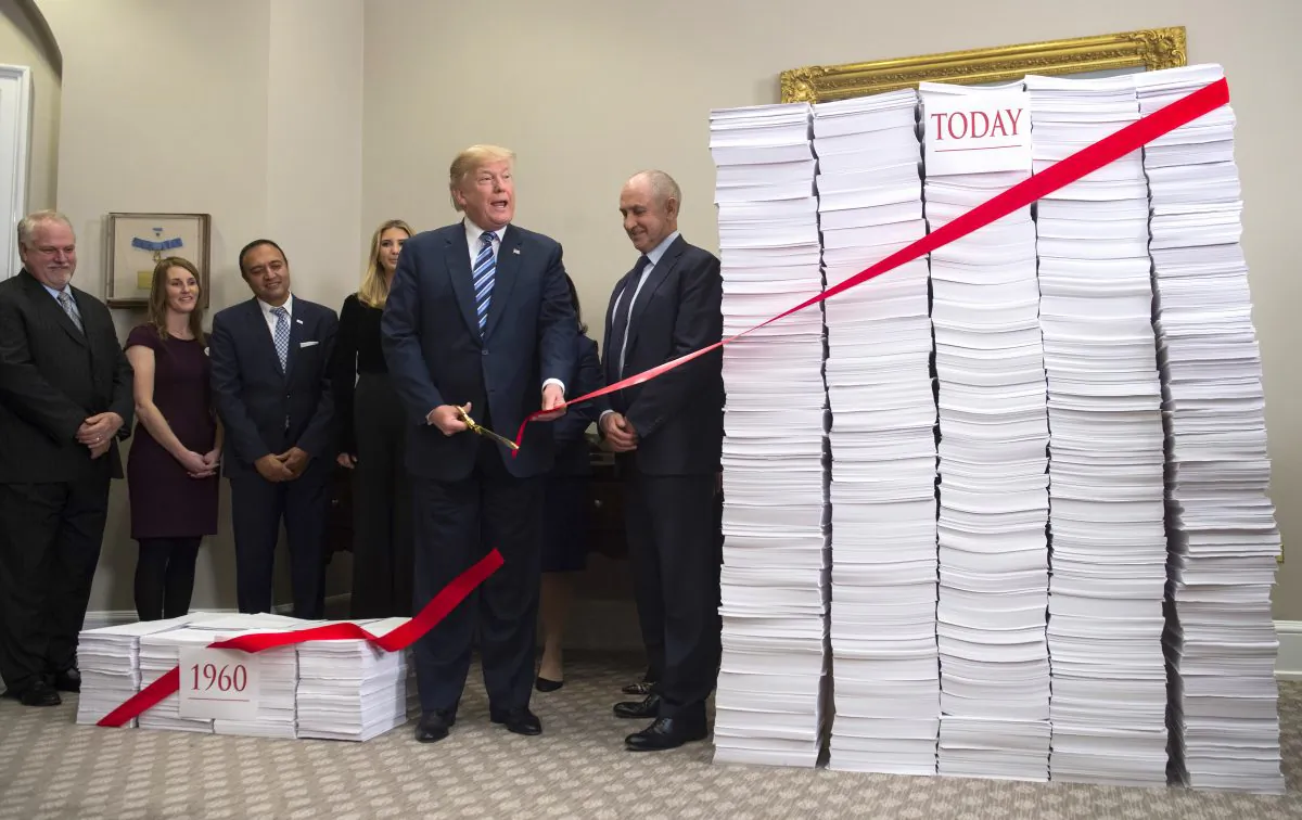 President Donald Trump uses a pair of golden scissors to cut red tape tied between two stacks of papers, representing the government regulations of the 1960s (L) and the regulations of today (R), in the Roosevelt Room of the White House on Dec. 14, 2017. (Saul Loeb/AFP/Getty Images)