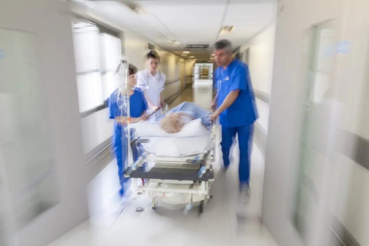 A motion blurred photograph of a patient on stretcher or gurney being pushed at speed through a hospital corridor by doctors & nurses to an emergency room (Pxhere)