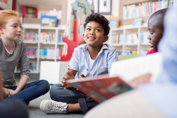Multiethnic group of kids sitting in circle around the teacher and listening a story. Discussion group of children inaa library talking to woman.