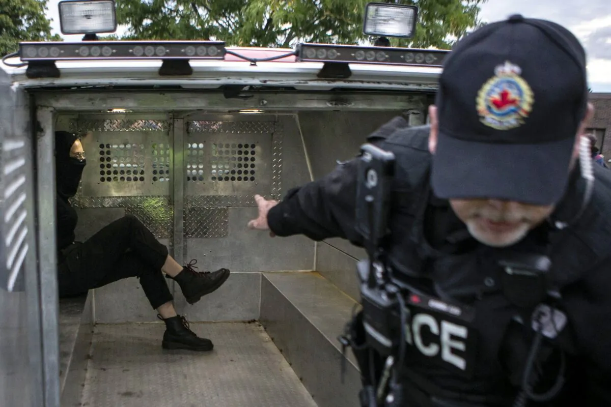 A black-clad masked man sits in a police van after being arrested at a demonstration prior to a  People's Party of Canada event in Hamilton, Ont., on Sept. 29, 2019. Three people have been charged in connection with a protest outside the event featuring PPC Leader Maxime Bernier, including a man whose family temporarily shut down their business amid backlash over his participation. (THE CANADIAN PRESS/Chris Young)
