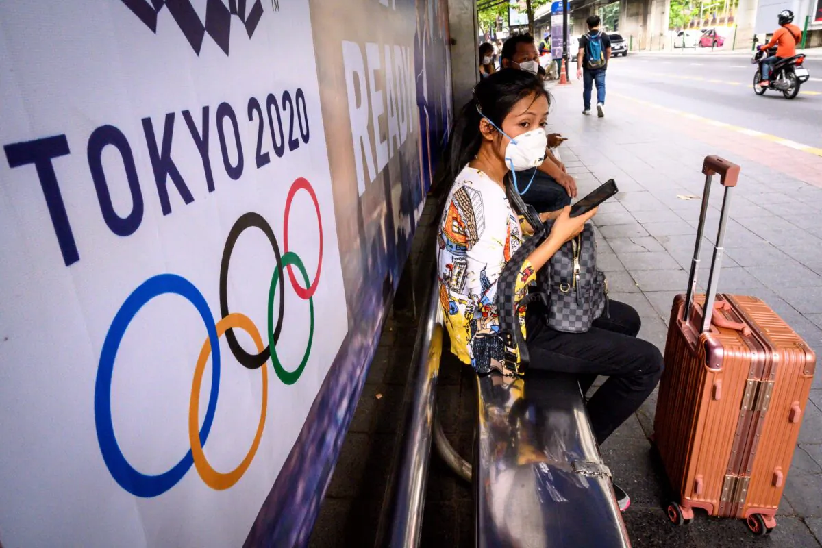 A woman with a suitcase wearing a facemask, amid concerns over the spread of COVID-19, sits at a bus stop in front of a Tokyo 2020 Olympics advertisement in Bangkok, Thailand, on March 16, 2020. (Mladen Antonov/AFP via Getty Images)