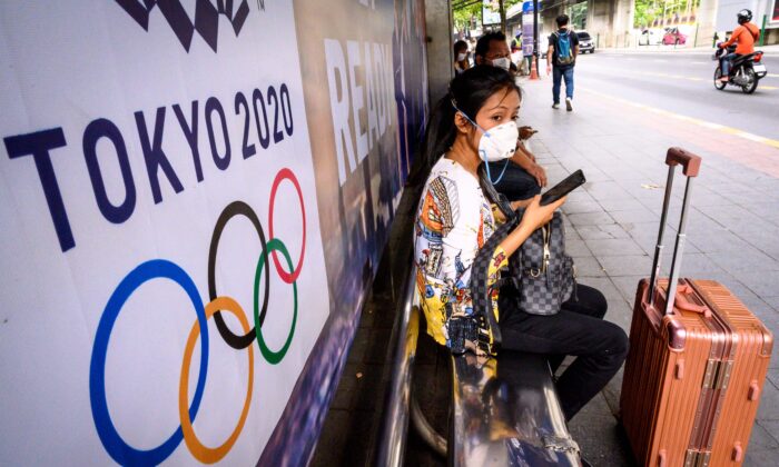 A woman with a suitcase wearing a facemask, amid concerns over the spread of COVID-19, sits at a bus stop in front of a Tokyo 2020 Olympics  advertisement in Bangkok, Thailand, on March 16, 2020. (Mladen  Antonov/AFP via Getty Images)