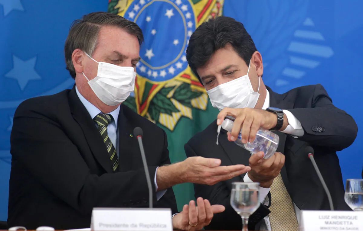 Brazilian Health Minister Luiz Henrique Mandetta (R) gives gel alcohol to President Jair Bolsonaro, both using protective masks, during a press conference about government plans and measures about the CCP virus crisis in Brazil, at the Planalto Palace in Brasilia, Brazil, on March 18, 2020. (Andre Coelho/Getty Images)