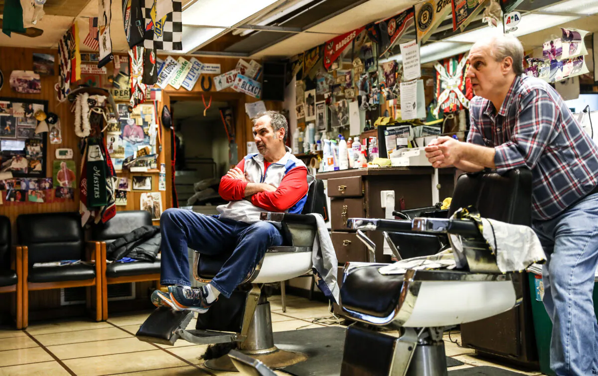 Barbers and brothers Tommy (L) and Jimmy Checkler watch the latest news on the coronavirus in their empty barbershop in the City of Worthington in Columbus, Ohio, on March 17, 2020. (Charlotte Cuthbertson/The Epoch Times)