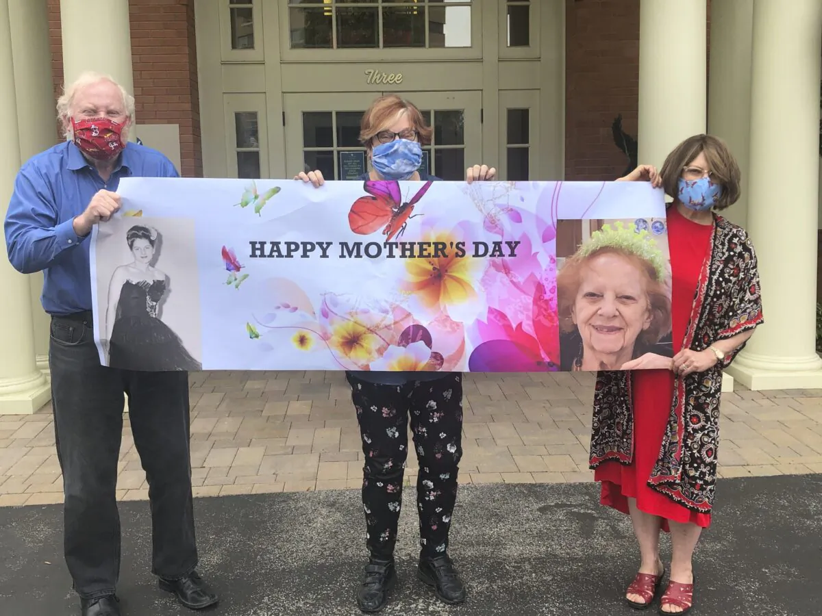From left, Steve Turner and his sisters, Carla Paull and Lisa Fishman, hold up a Mother’s Day banner emblazoned with images of their mom, Beverly Turner, in front of her assisted living facility in Ladue, Mo., on May 3, 2020. (Shelly Solomon via AP)