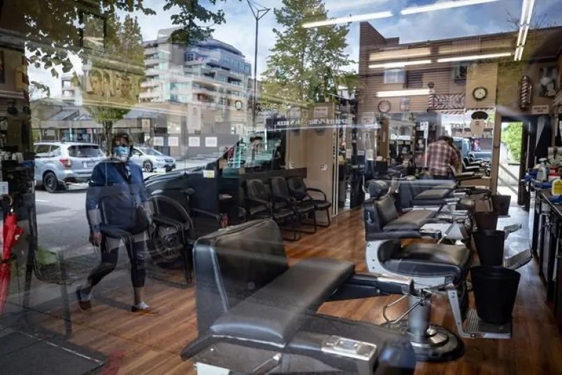 A man cleans inside a closed barbershop as a pedestrian wearing a face mask is reflected in the shop's window, in Burnaby, B.C., on May 6, 2020. (Darryl Dyck/The Canadian Press) 