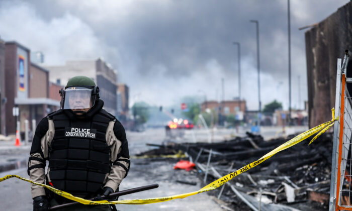 A police officer stands amid smoke and debris as buildings continue to burn in the aftermath of a night of protests and violence following the death of George Floyd, in Minneapolis, Minn., on May 29, 2020. (Charlotte Cuthbertson/The Epoch Times)