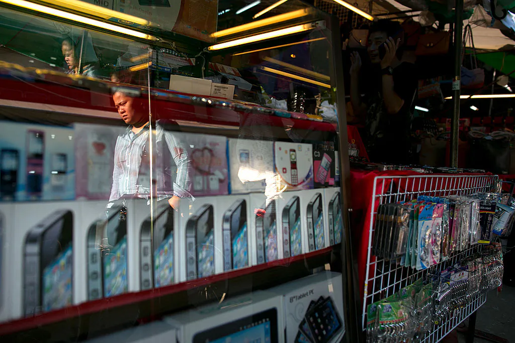 A man is reflected in the glass of display case in a shop selling counterfeit Chinese mobile phones at an outdoor market in the Golden Triangle, situated along the Thai- Burma border November 12, 2012 in Tachiliek, Myanmar. (Paula Bronstein/Getty Images)