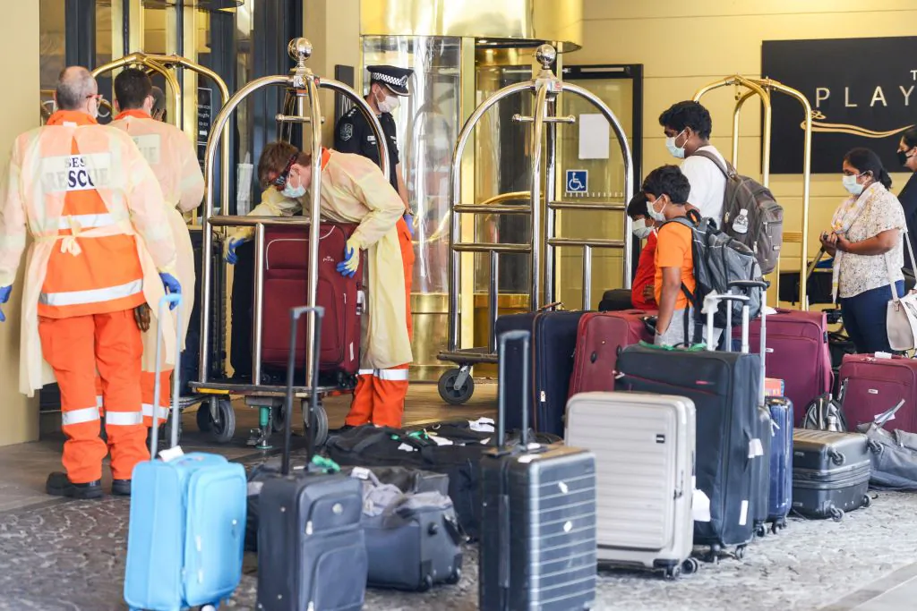 Local officials (L) in protective gear help with baggage as Australian residents (R) arrive at a hotel in Adelaide for mandatory 14-day quarantine after returning from overseas on April 21, 2020. (Brenton EDWARDS / AFP via Getty Images)
