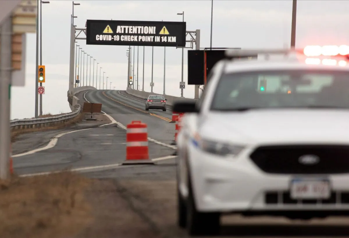 An RCMP vehicle in front of the Confederation Bridge as a vehicle passes under a warning sign of the coronavirus disease (COVID-19) checkpoint on Prince Edward Island at Cape Jourimain, New Brunswick, Canada, on April 2, 2020. (Reuters/John Morris)